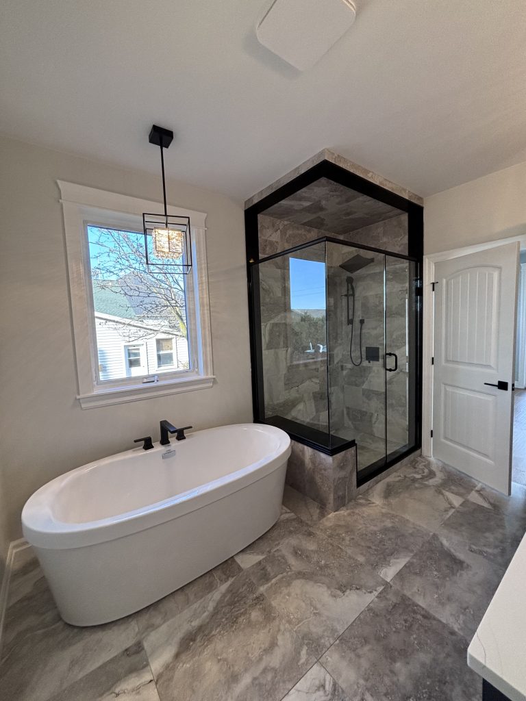 Modern bathroom with a freestanding white tub beside a glass shower enclosure and marble tile floor. A square pendant light hangs near a window.