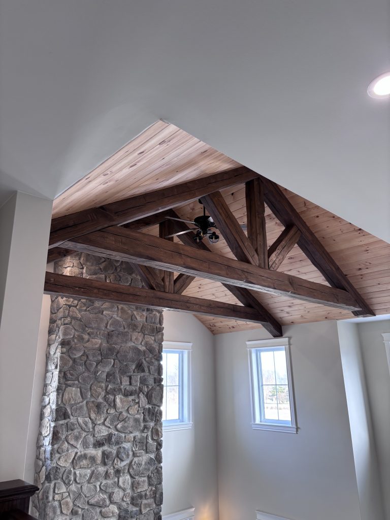 Interior view featuring a tall stone fireplace and dark wooden exposed beams under a vaulted wooden ceiling with a ceiling fan and windows letting in light.