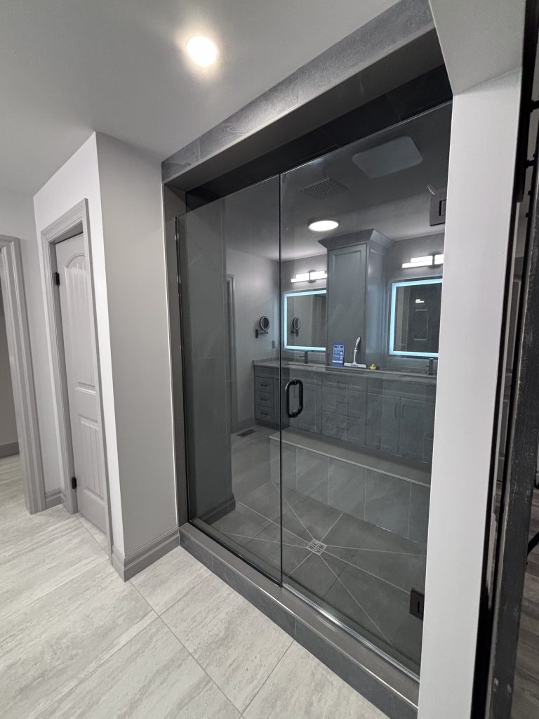 Glass-walled shower enclosure in a modern bathroom with gray tile and a vanity reflected in the glass