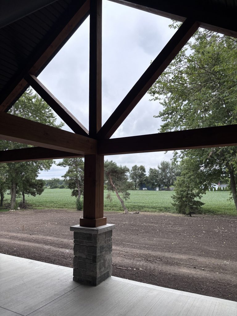 Wooden pavilion frame with diagonal beams seen from inside, overlooking a grassy field and trees.